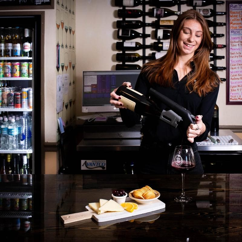 A smiling woman is pouring wine into a glass at a bar, with a cheese platter and snacks in front of her.