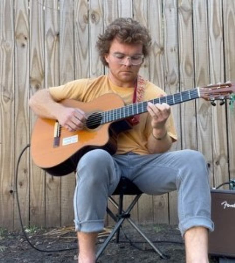 A person with curly hair sits on a chair playing an acoustic guitar outdoors against a wooden fence.