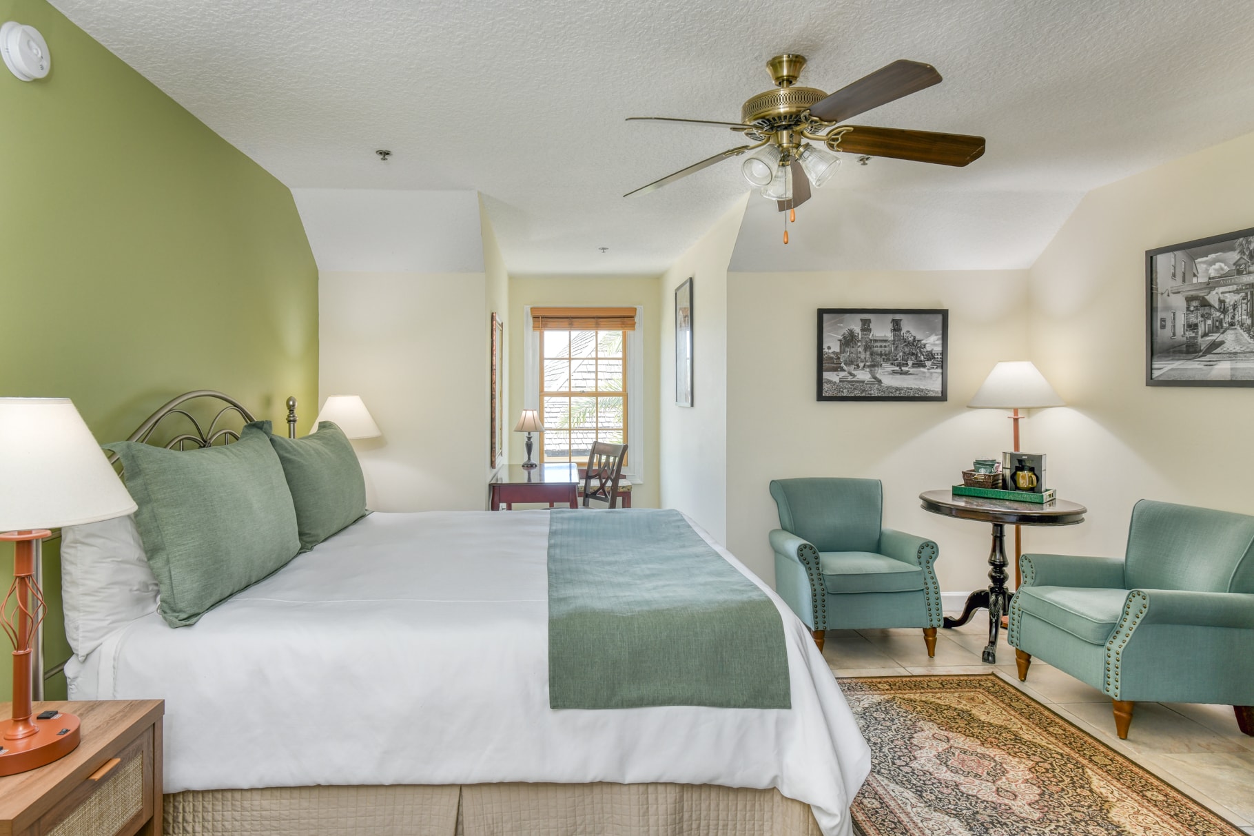 A cozy hotel room featuring a bed, seating area, and a small table, with light green walls and natural light coming through the window.