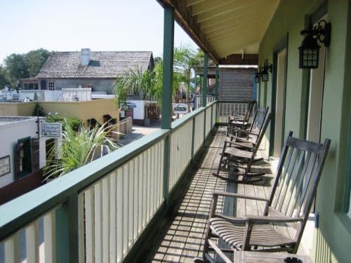 A sunlit balcony with rocking chairs overlooks a quaint street lined with shops and palm trees.
