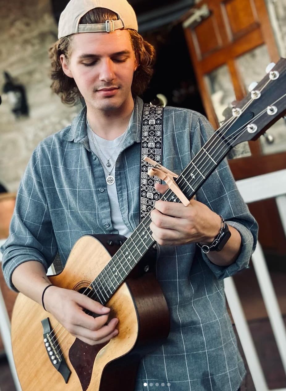 A young man in a cap plays an acoustic guitar while seated, focusing intently on his music.