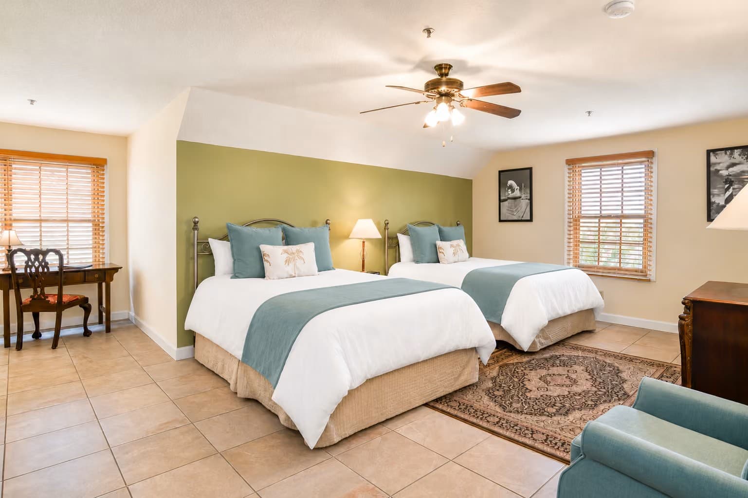 A cozy hotel room featuring two beds, a desk, and natural light from windows.