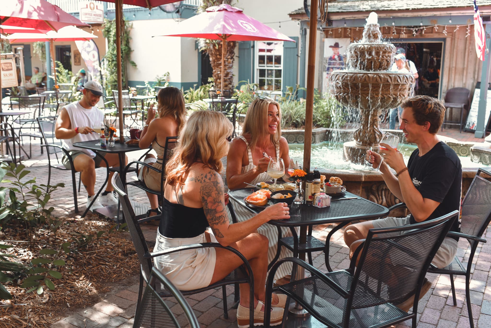 A group of friends enjoys meals and drinks at an outdoor café near a fountain.