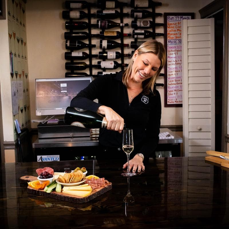 A woman pours champagne into a glass next to a charcuterie board in a wine bar setting.