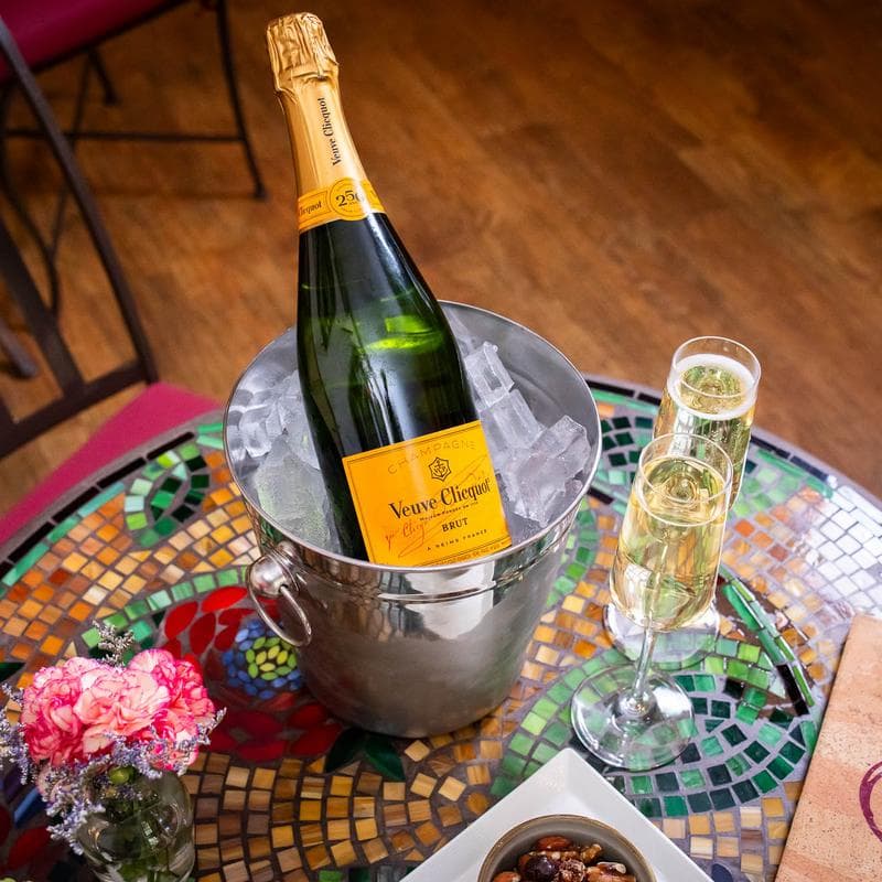 A bottle of Veuve Clicquot champagne in an ice bucket next to two glasses of champagne and a bowl of nuts on a colorful table.