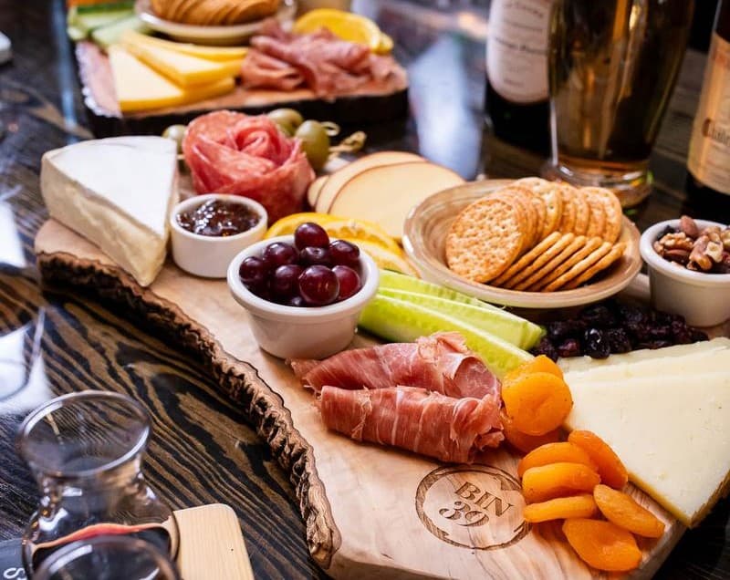 A wooden platter featuring an assortment of cheeses, cured meats, crackers, grapes, and dried fruits.