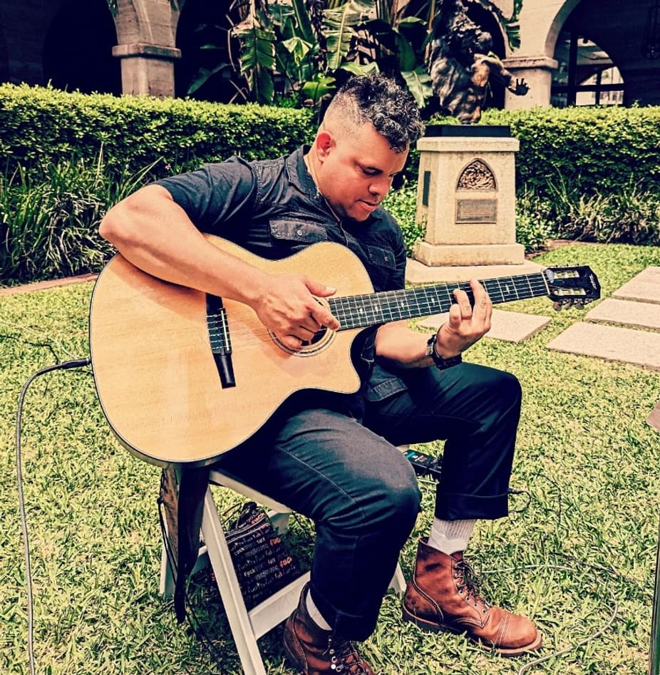A musician sits on a chair playing an acoustic guitar in a grassy area surrounded by plants.