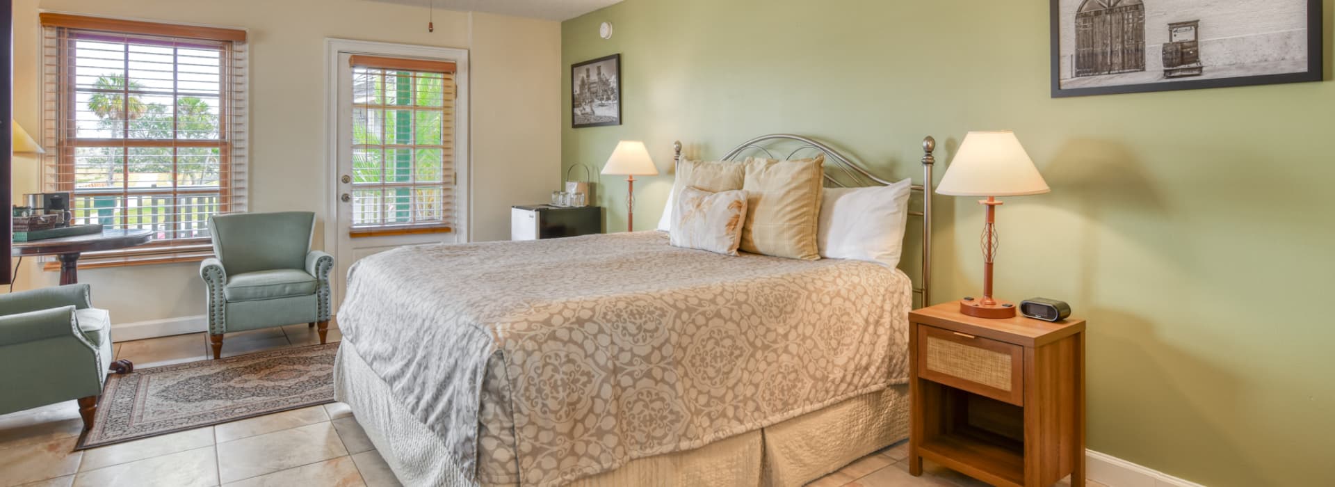 A cozy bedroom featuring a bed, armchairs, a side table with a lamp, and large windows.