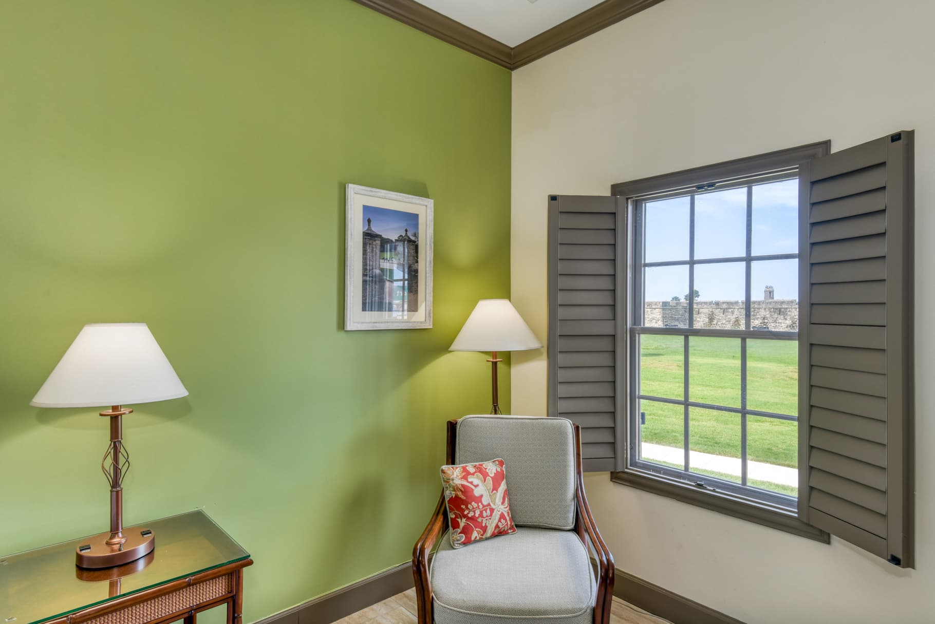 A cozy corner with a chair, lamp, and a window showing a green landscape, against a green wall.
