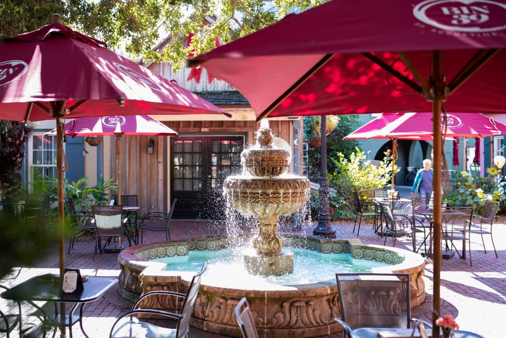 A decorative fountain surrounded by tables and red umbrellas in a quaint outdoor seating area.