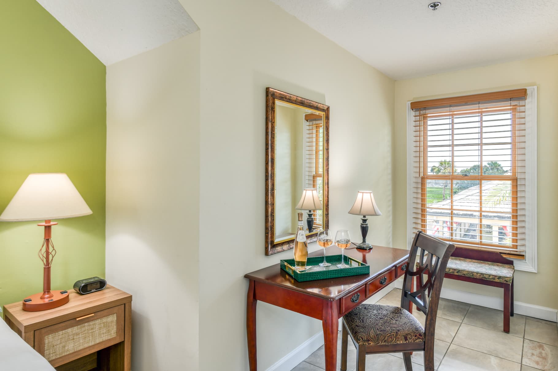 A cozy corner in a room featuring a desk with a mirror, lamps, and a view through a window.