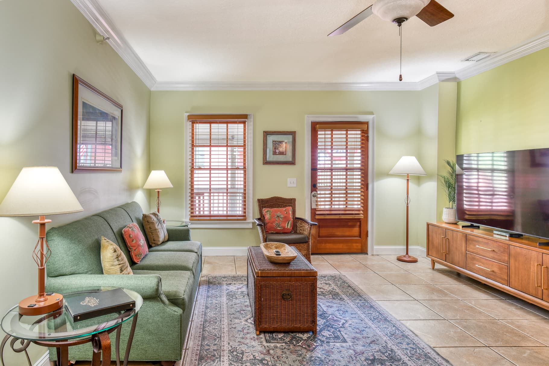 A cozy living room with green walls, a sofa, a wooden coffee table, a television, and natural light from window shutters.