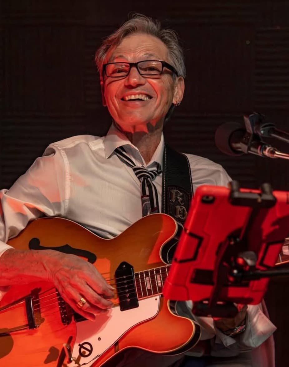 A smiling musician in a white shirt and tie plays an orange guitar on stage.