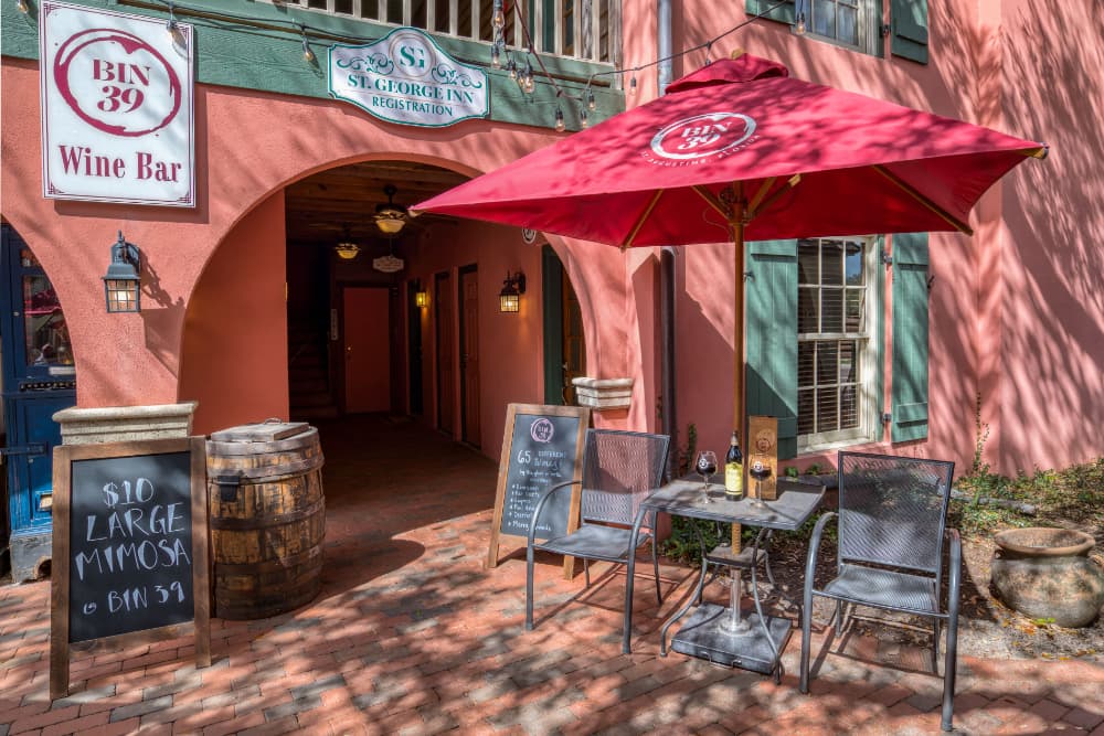 Outdoor seating with a red umbrella at a wine bar entrance featuring signs and a chalkboard menu.