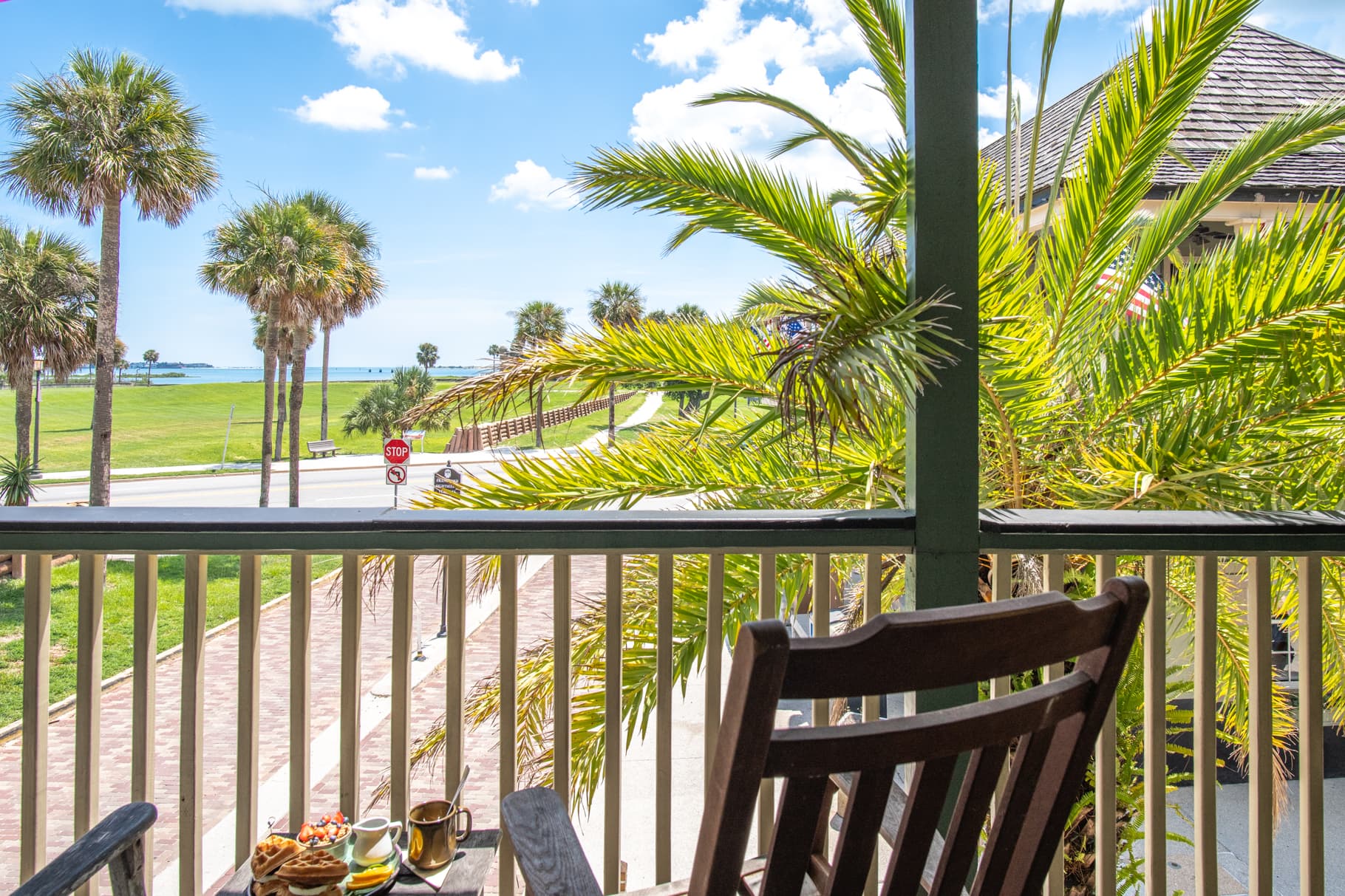 View from a balcony with palm trees overlooking a road and the ocean under a blue sky.