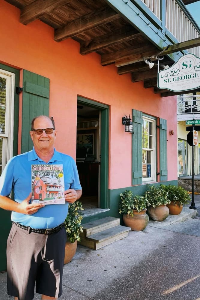 A smiling man in a blue shirt holds a magazine in front of a pink building with a sign that reads "St. George."