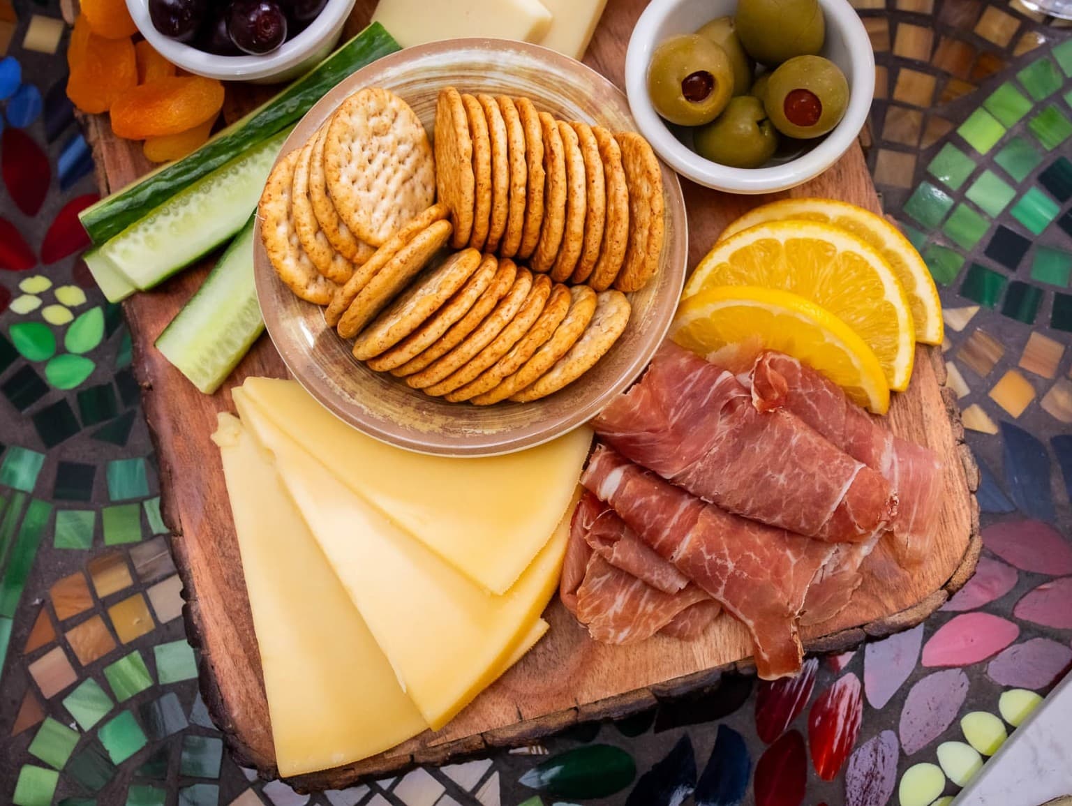 A wooden platter featuring crackers, cheese, cured meat, olives, cucumber, dried fruit, and orange slices on a colorful mosaic table.