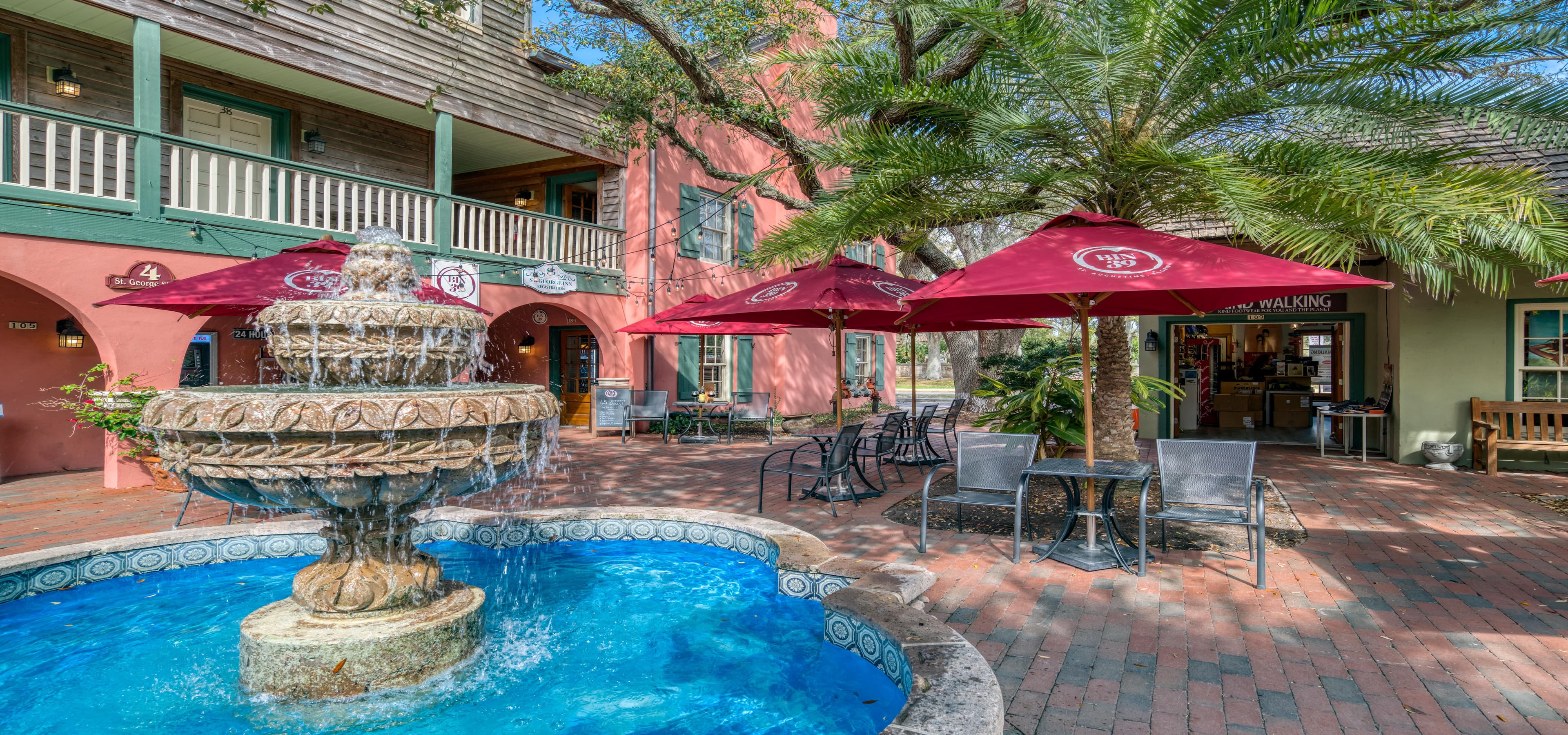 A charming courtyard featuring a fountain, surrounded by palm trees and café seating under red umbrellas.