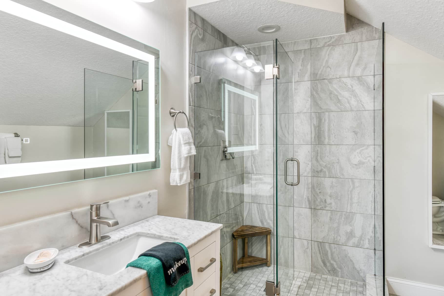 Modern bathroom with a glass shower, marble countertop, and illuminated mirror.