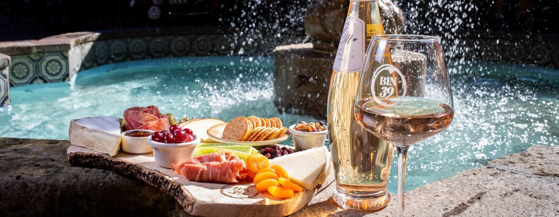 A charcuterie board with cheese, meats, and fruits beside a glass of wine and a bottle, set against a splashing fountain.