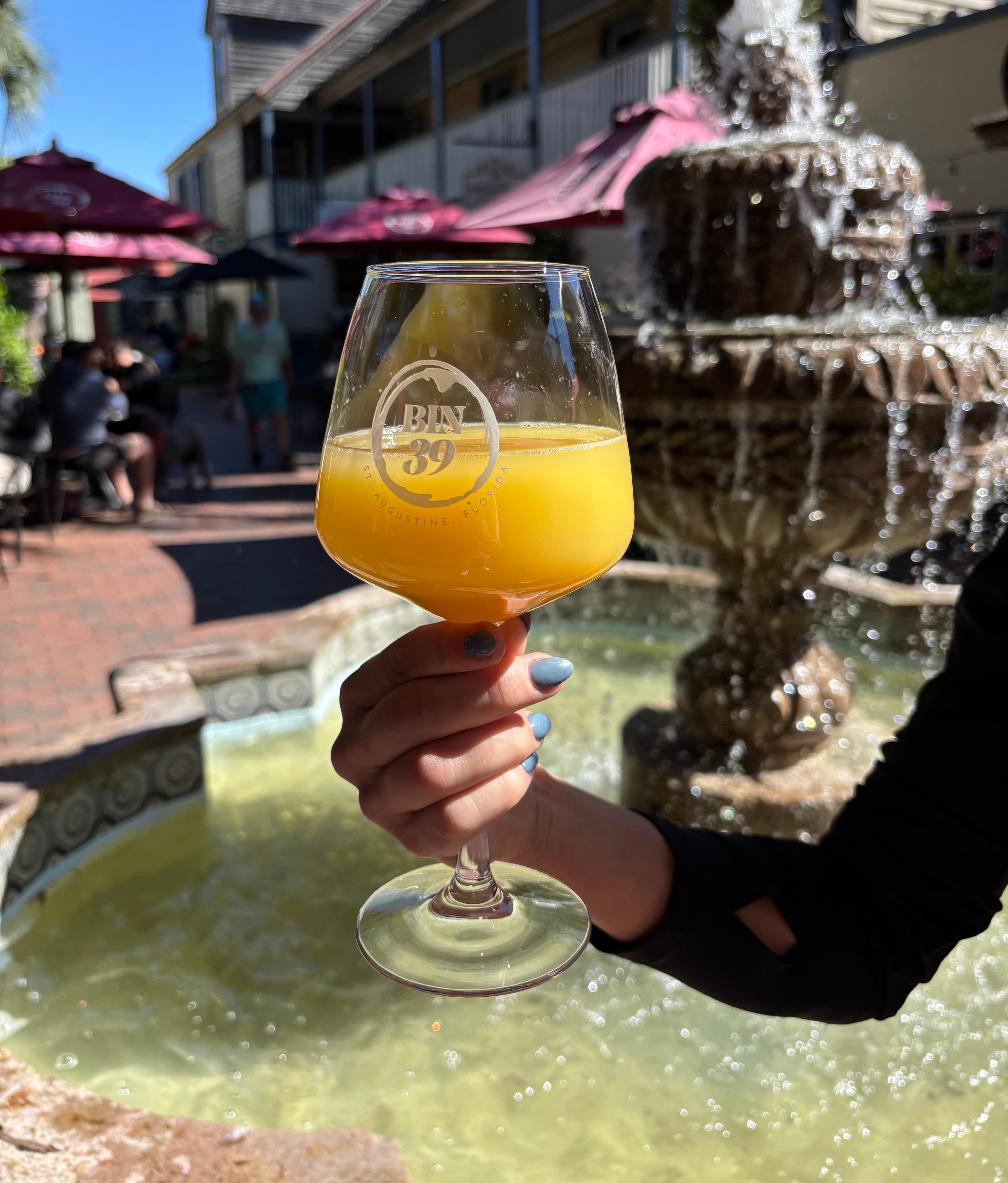 A hand holds a glass of orange juice in front of a fountain and outdoor café seating.