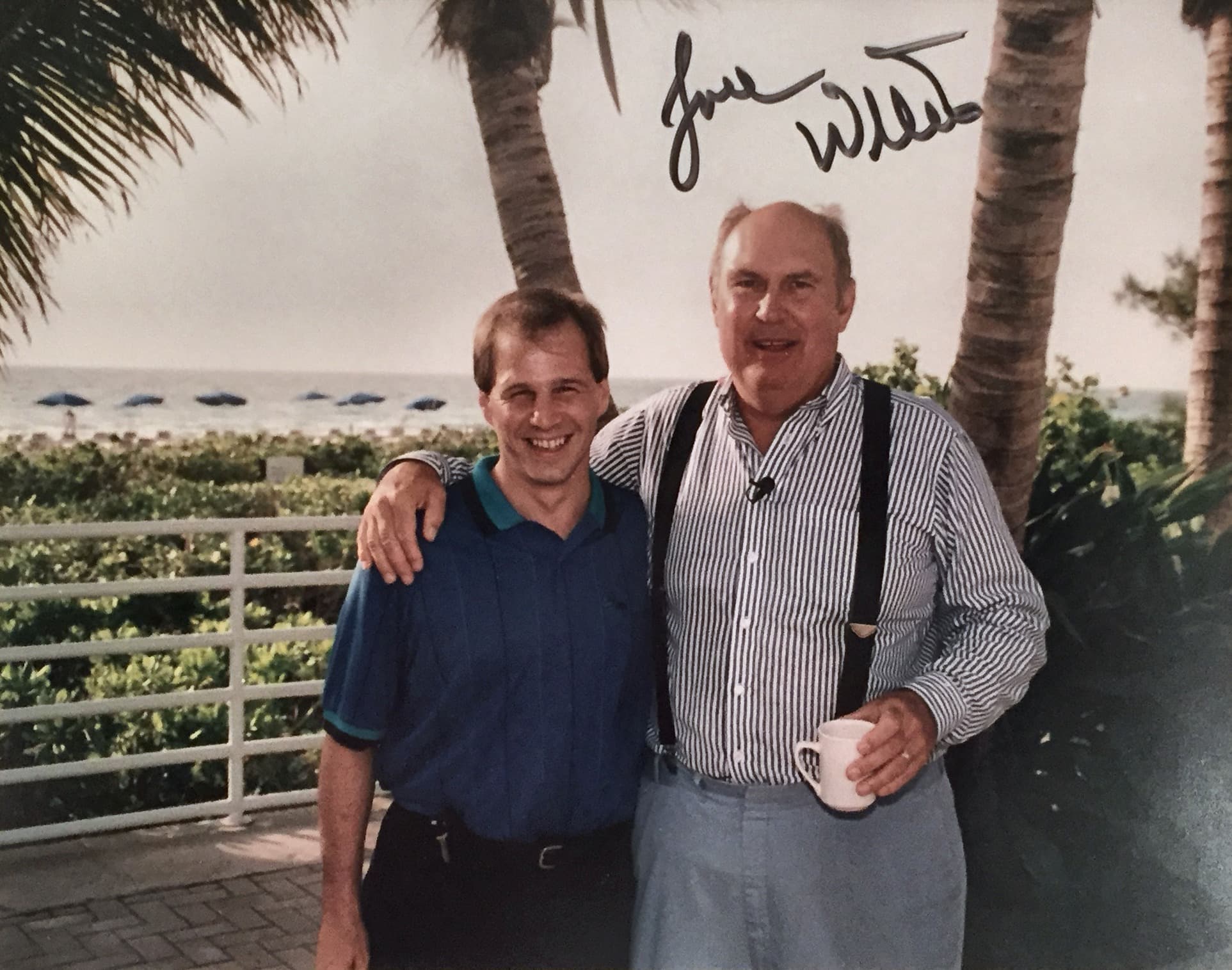 Two men stand together smiling in front of a beach, with palm trees in the background.