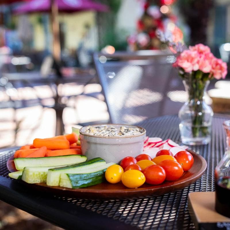 A wooden platter with various vegetables, cherry tomatoes, and a bowl of dip on a table outdoors.