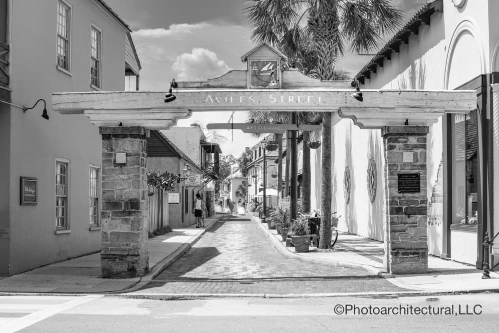 Black and white image of a quaint street with an archway labeled "Aviles Street" and cobblestone pathway lined with potted plants.