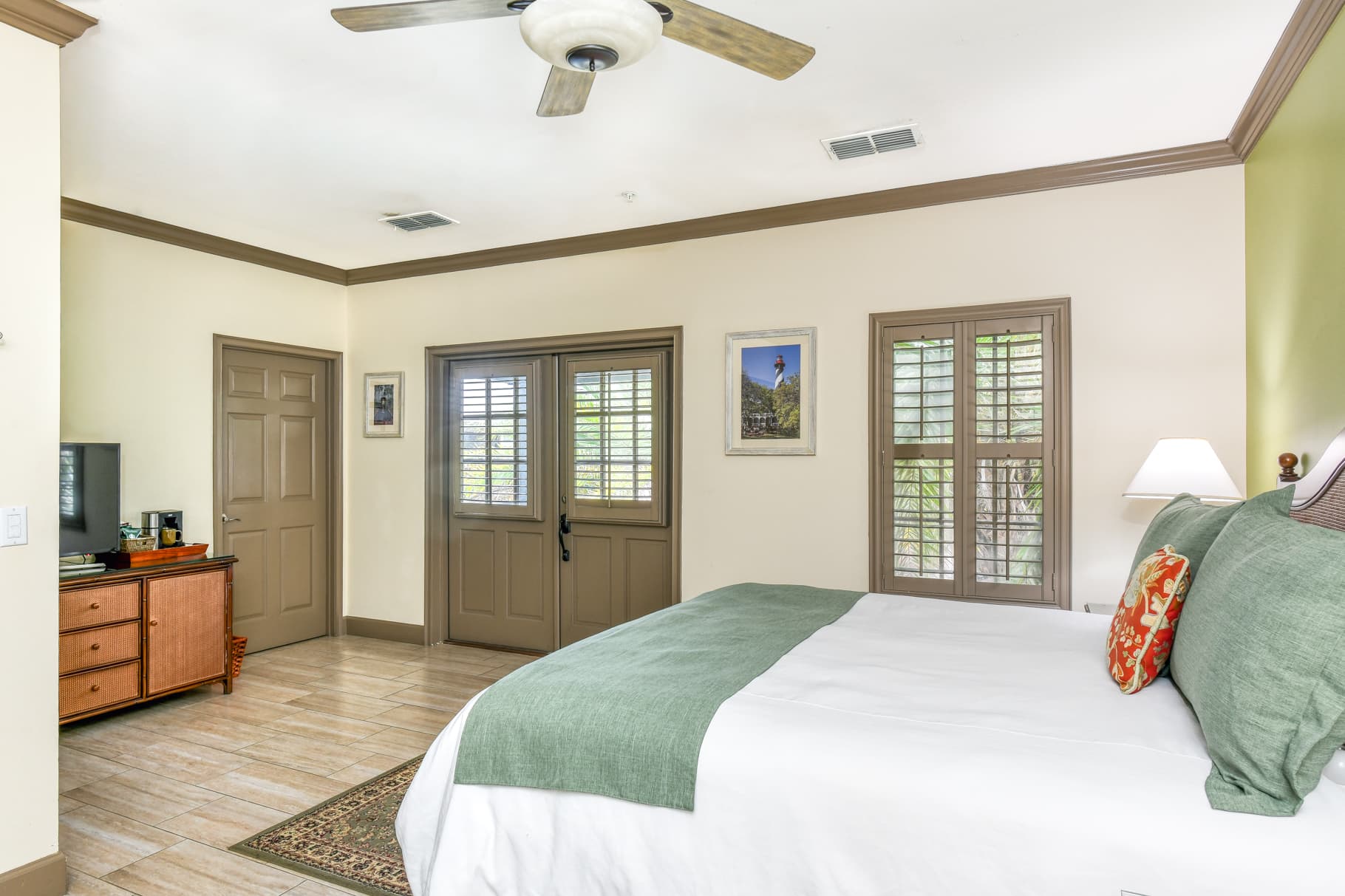 A well-lit bedroom featuring a queen-sized bed with green accents, a wooden dresser, a TV, and two sets of shutters on the windows.