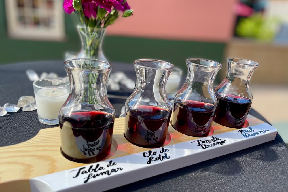 A wooden flight board holds four glass carafes of red liquid, accompanied by a small candle and flower arrangement on a table.