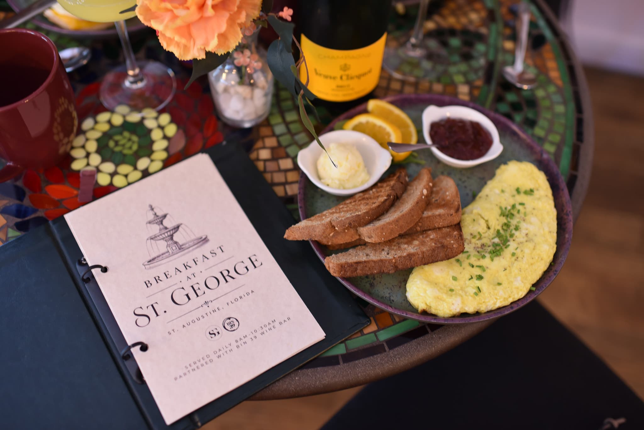 A plate of an omelet, toasted bread, cream, and orange slices next to a breakfast menu and a bottle of champagne on a colorful table.