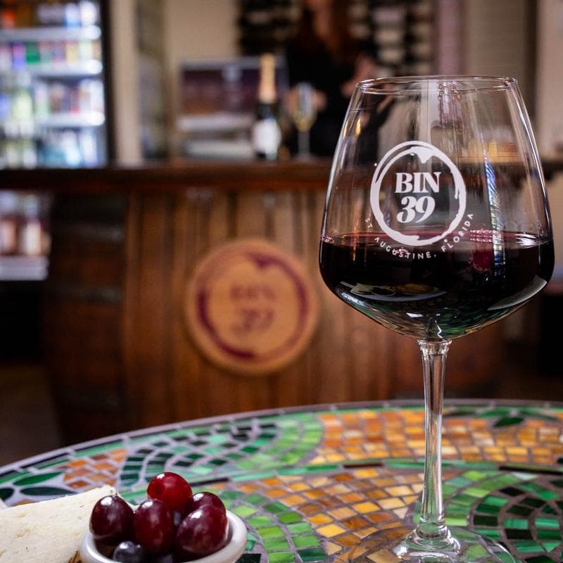 A glass of red wine sits on a colorful mosaic table with grapes and cheese, against a backdrop of a wine shop bar.
