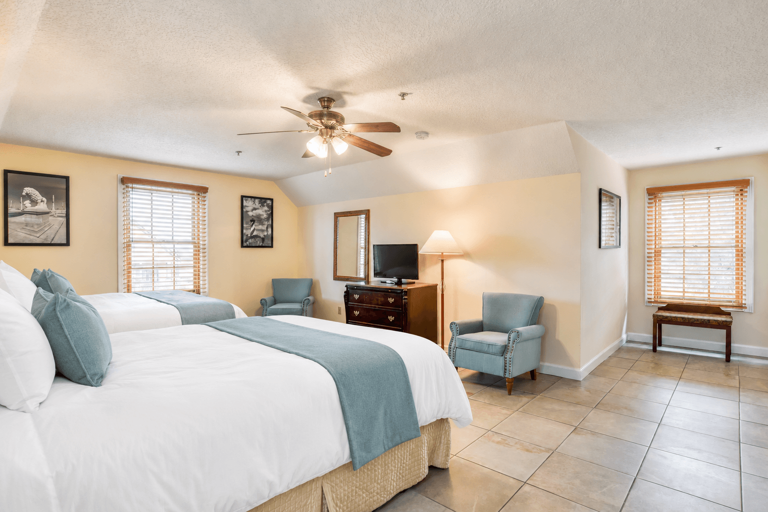 A spacious hotel room featuring two beds, a TV, and seating areas with natural light from windows.
