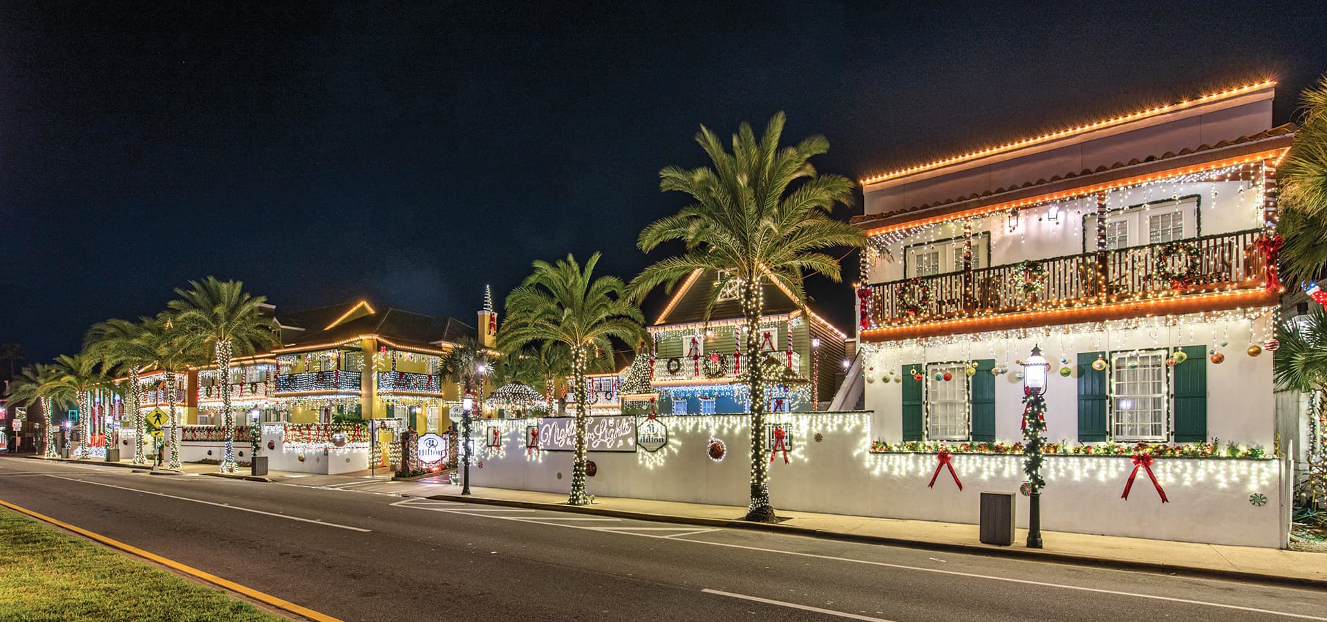 A row of brightly decorated houses with festive lights and palm trees, illuminated at night.